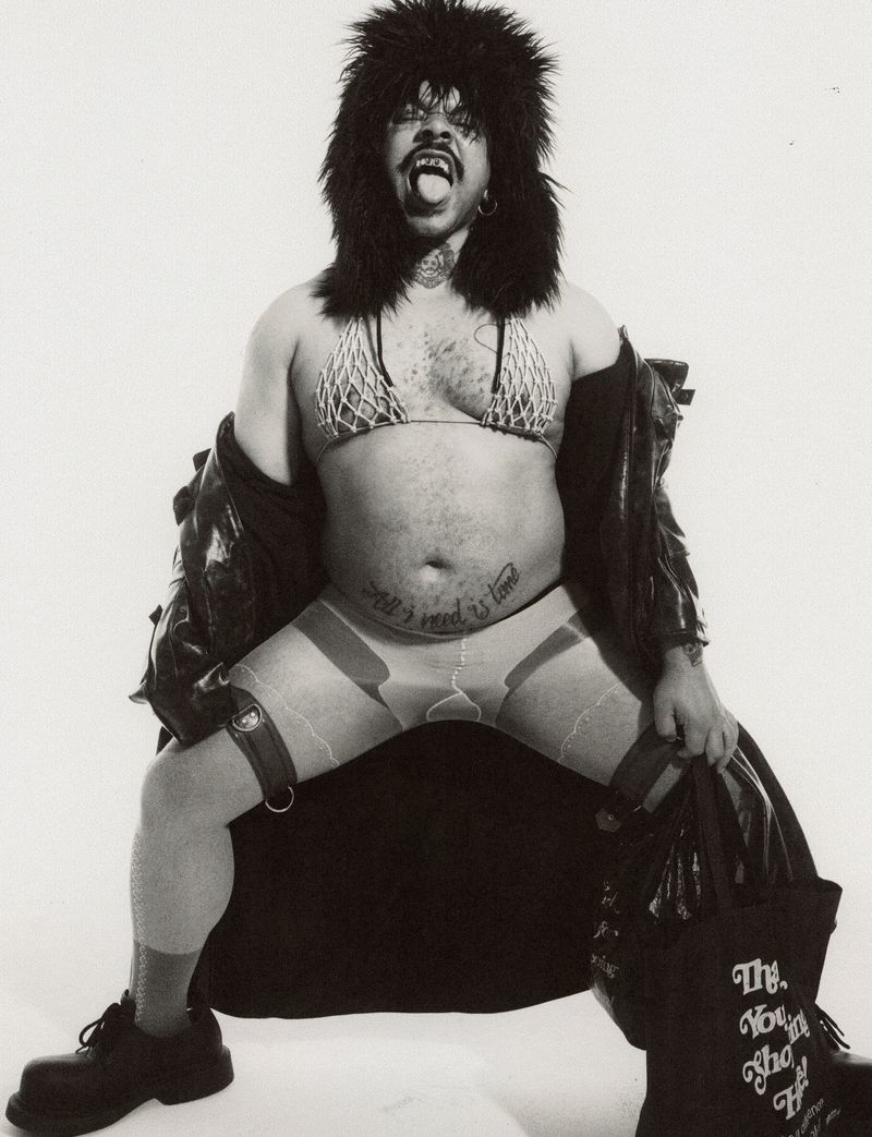 Monochrome portrait of a non-binary person wearing a fur hat and fishnet bikini top, with tattoos, holding a leather jacket and boots.
