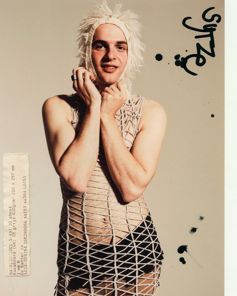 Person with shaggy blonde hair, wearing an open-weave top and skirt, posing in a photo studio against a neutral background with ink splatter.