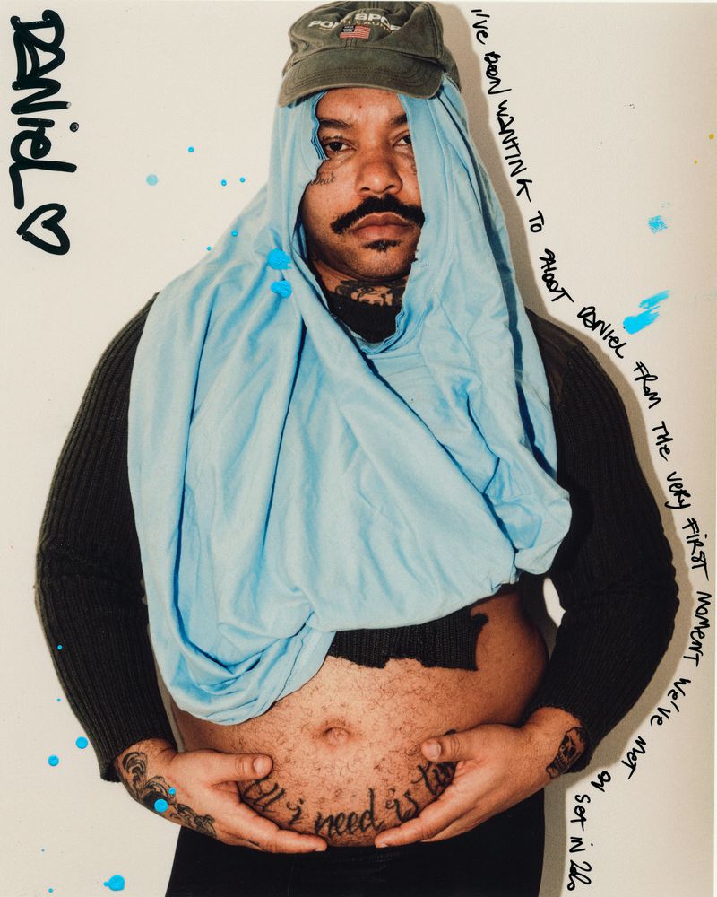 Portrait of an adult male with a mustache and tattoos, wearing a hat and light-blue fabric, against an off-white backdrop.