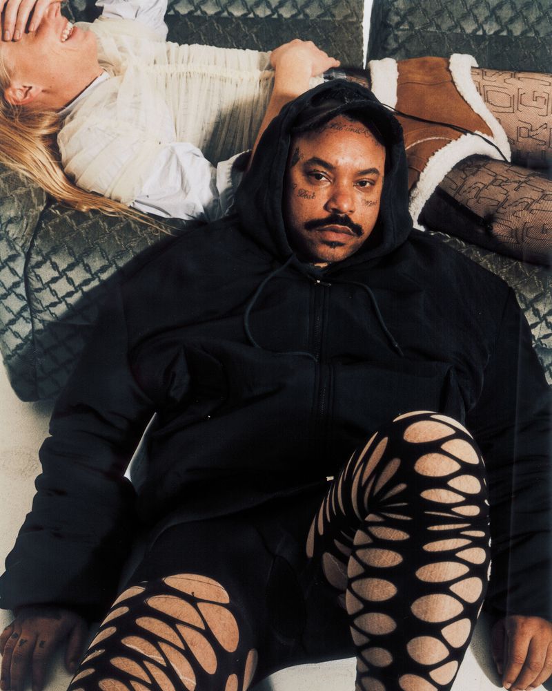Man in black hoodie and patterned tights poses with reclining woman on quilted bed. Fashion editorial with edgy, urban aesthetic.