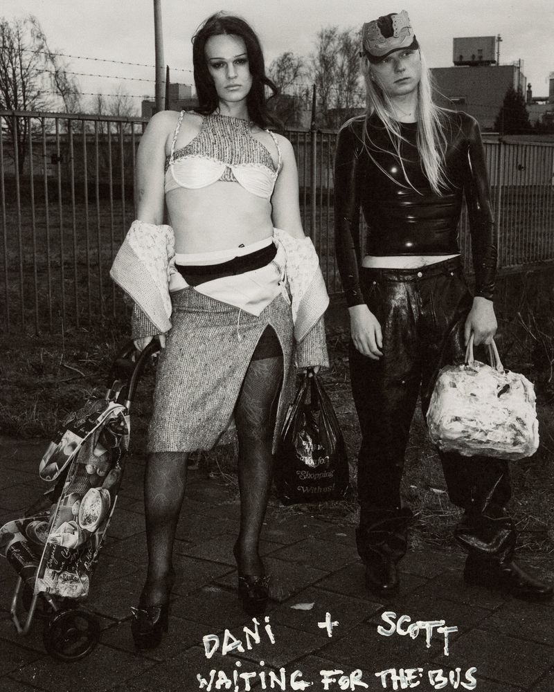 Two adults in black and white urban outdoor setting; woman in skirt and bra with shopping cart, man in latex top and cap holds bag.