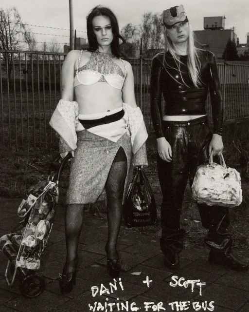 Two adults in black and white urban outdoor setting; woman in skirt and bra with shopping cart, man in latex top and cap holds bag.