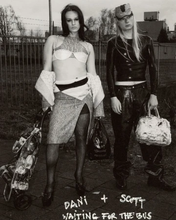 Two adults in black and white urban outdoor setting; woman in skirt and bra with shopping cart, man in latex top and cap holds bag.