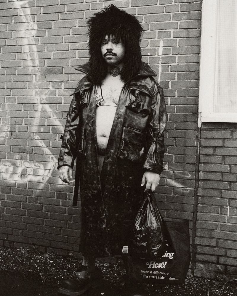 Monochrome portrait of a person in a fur hat and black coat holding a shopping bag, standing against a brick wall outdoors.