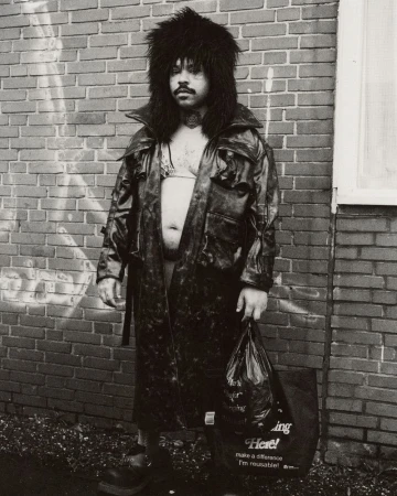 Monochrome portrait of a person in a fur hat and black coat holding a shopping bag, standing against a brick wall outdoors.