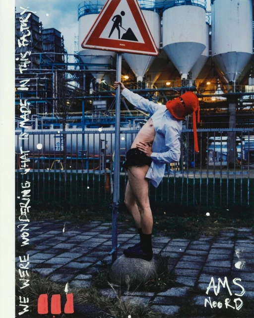 An adult with an orange hood stands on a rock next to a road work sign in front of a large industrial plant on a cloudy day.