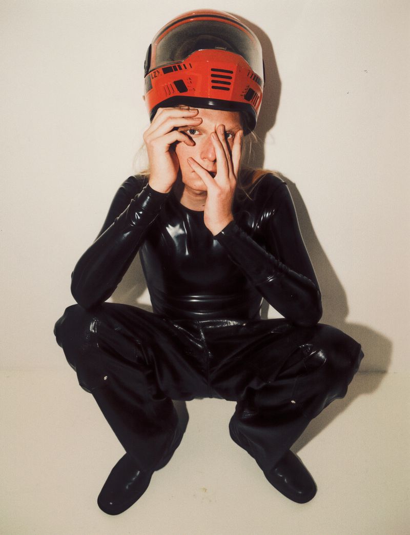 Person in a black catsuit and red helmet kneels in front of a plain backdrop with hands near their face, creating a striking and unusual portrait.