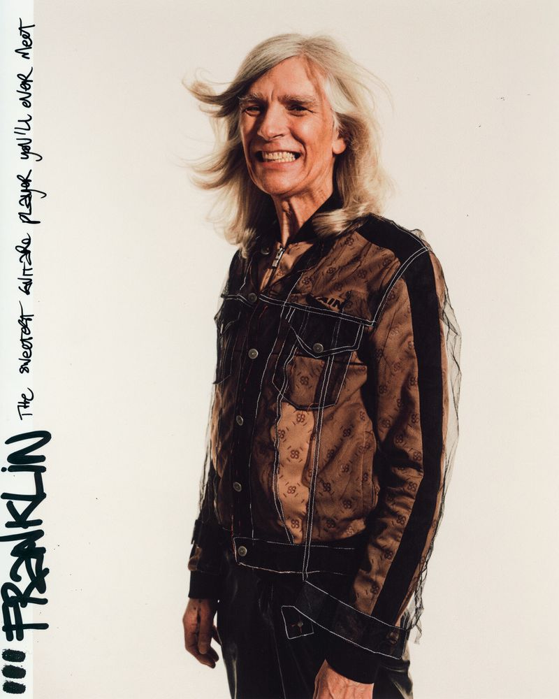 Portrait of a smiling adult man with long blond hair wearing a brown patterned jacket against a bright white background in a studio.