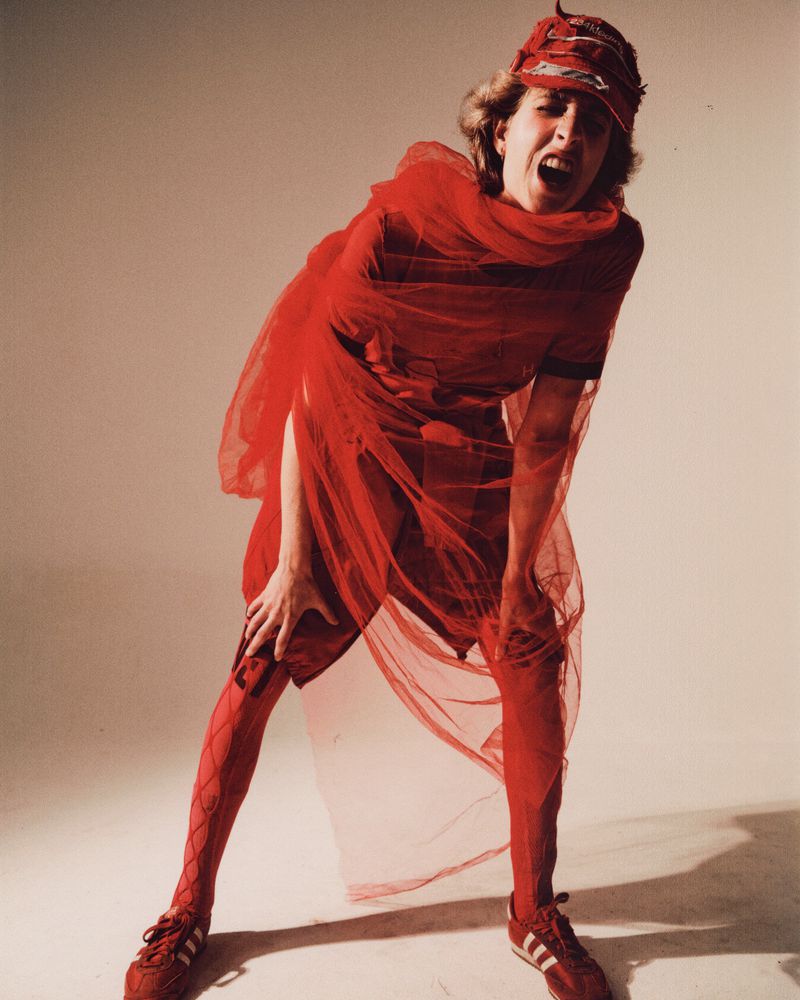 Woman in red tulle fabric and Adidas sneakers strikes pose with hands on knees and an open mouth in a photo studio setting.