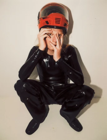 Model wearing a black glossy bodysuit and red motorcycle helmet squatting against a plain white wall