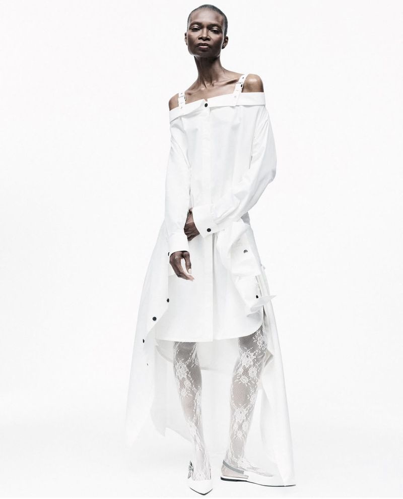 Fashion model posing in a white, off-the-shoulder dress with lace leggings in a studio setting, showcasing a unique style.