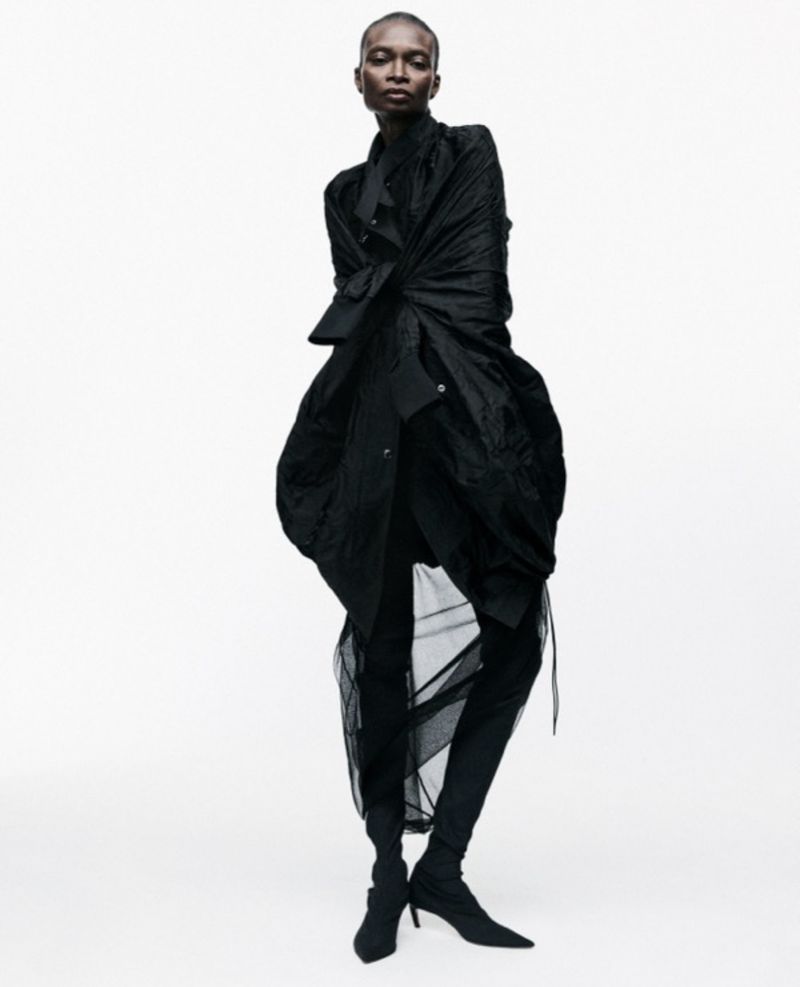 An adult female poses in a full-length studio shot, wearing a draped black coat, leggings, and ankle boots against a white background.