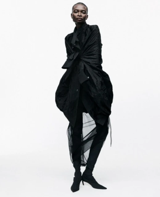 An adult female poses in a full-length studio shot, wearing a draped black coat, leggings, and ankle boots against a white background.