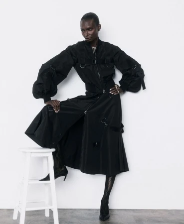 Model Debra Shaw in a black jacket and long skirt posing with a white stool on a white background