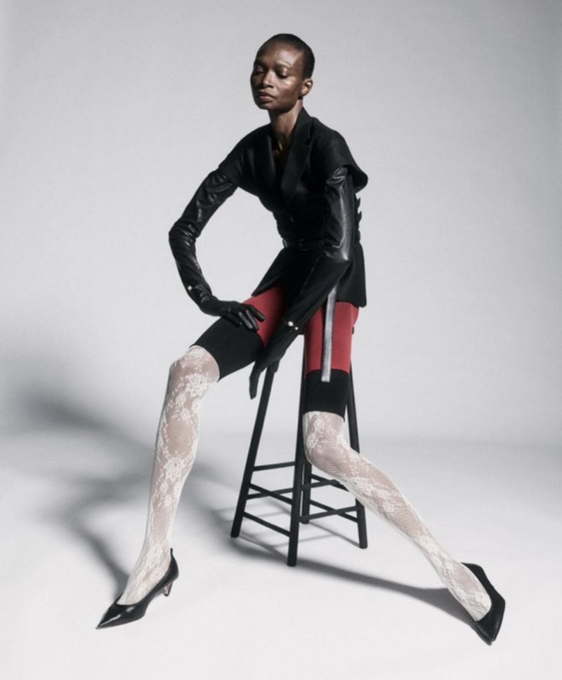 Model Debra Shaw seated on a stool wearing a black blazer, red shorts, lace tights and black heels against a light grey background