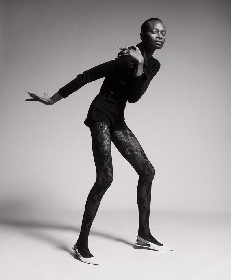 Black and white portrait of an African American woman in a black outfit and lace tights, striking a pose against a neutral backdrop.