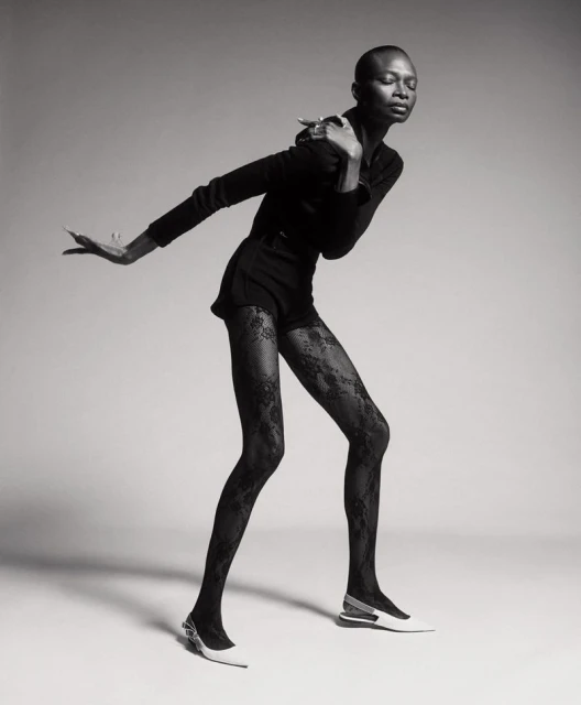 Black and white portrait of an African American woman in a black outfit and lace tights, striking a pose against a neutral backdrop.