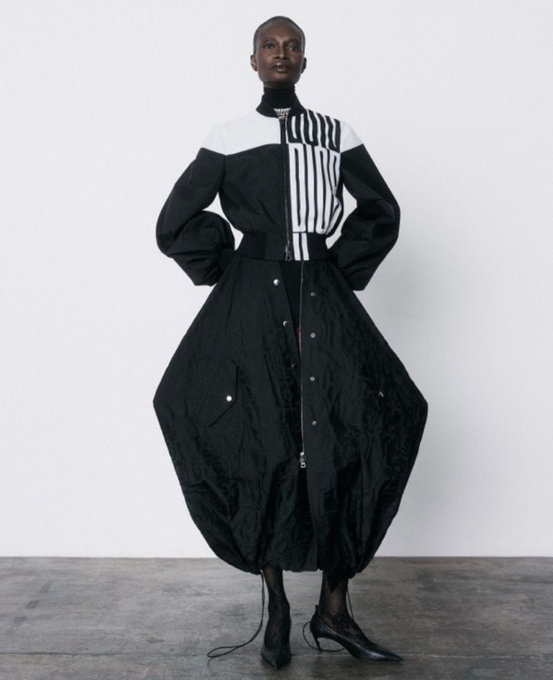 Striking studio shot of a black adult woman modeling a black and white jacket with a voluminous black skirt and heeled boots.