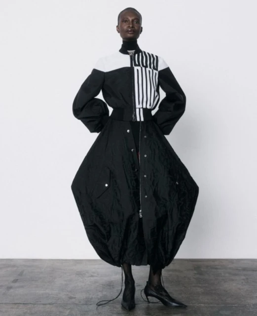 Striking studio shot of a black adult woman modeling a black and white jacket with a voluminous black skirt and heeled boots.