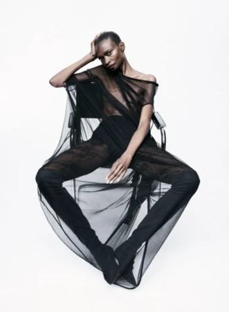 Model Debra Shaw seated on a modern chair in a sheer black draped garment against a plain white background