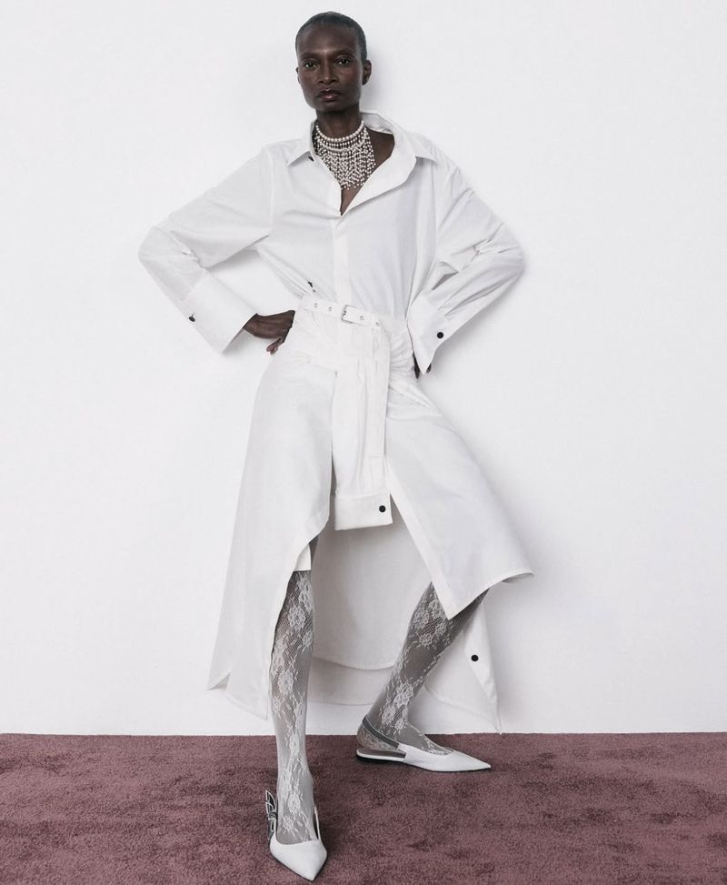 A slender Black female fashion model poses in a long white belted shirt-dress, stockings, pumps, and beaded necklace against a white wall.