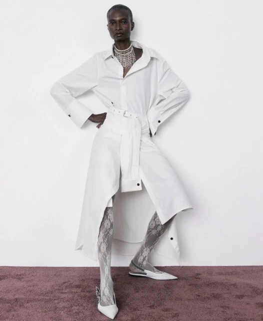 A slender Black female fashion model poses in a long white belted shirt-dress, stockings, pumps, and beaded necklace against a white wall.