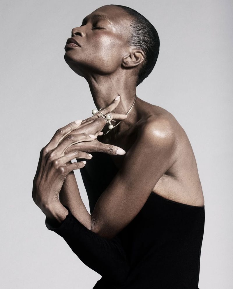 Elegant portrait of a Black woman in a black top, posing with hands near her neck, wearing jewelry against a neutral backdrop.