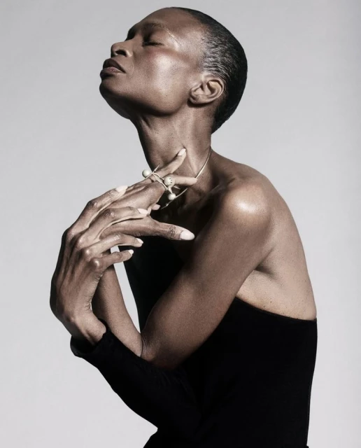 Elegant portrait of a Black woman in a black top, posing with hands near her neck, wearing jewelry against a neutral backdrop.