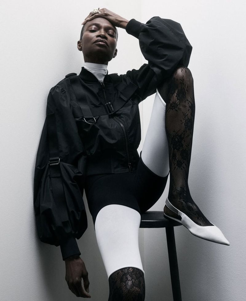 Elegant, dark-skinned fashion model poses leaning against a wall, wearing a black jacket, black and white shorts, lace stockings, and white shoes.