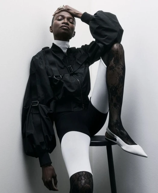 Elegant, dark-skinned fashion model poses leaning against a wall, wearing a black jacket, black and white shorts, lace stockings, and white shoes.