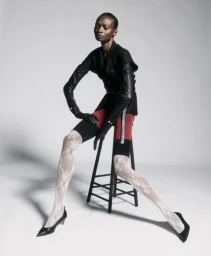 Model Debra Shaw seated on a stool wearing a black blazer, red shorts, lace tights and black heels against a light grey background