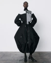 Model Debra Shaw wearing a sculptural black and white bomber jacket and voluminous skirt against a plain white studio backdrop.