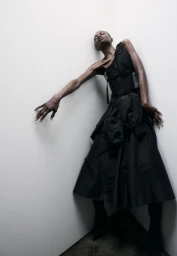 Model in black utility vest and flared skirt leaning against a white studio corner wall