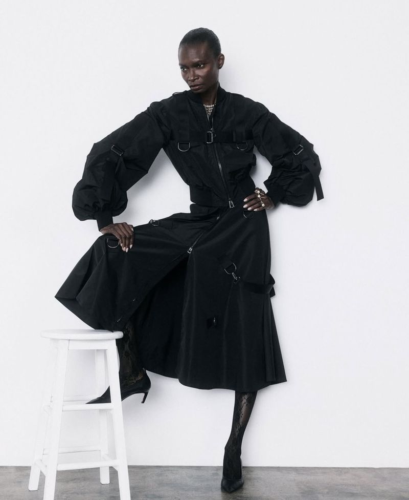 Model Debra Shaw in a black jacket and long skirt posing with a white stool on a white background