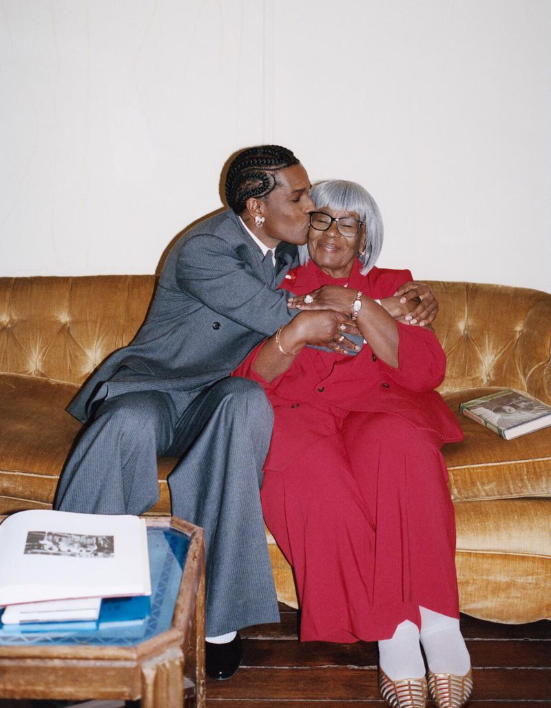 Rapper A$AP Rocky embraces and kisses his elderly grandma on the cheek while sitting on a couch, with warm indoor lighting.