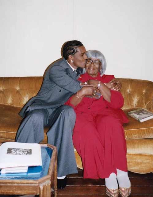 Rapper A$AP Rocky embraces and kisses his elderly grandma on the cheek while sitting on a couch, with warm indoor lighting.