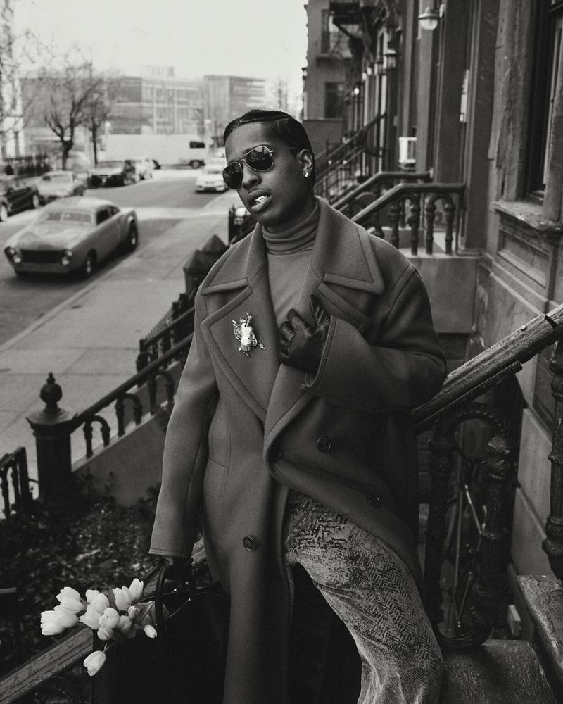 Monochrome shot of stylish man in overcoat, sunglasses, and gloves standing on stairs outdoors with flowers and buildings.