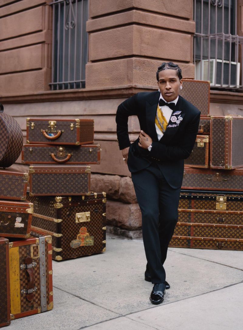 Stylish adult male in a black tuxedo suit poses next to stacked vintage Louis Vuitton suitcases in an urban setting with tan building.