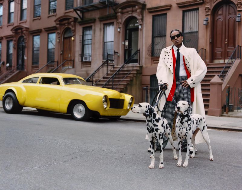 Stylish man in a polka dot outfit walks two Dalmatians on leashes in front of a yellow classic car and brownstones in an urban setting.