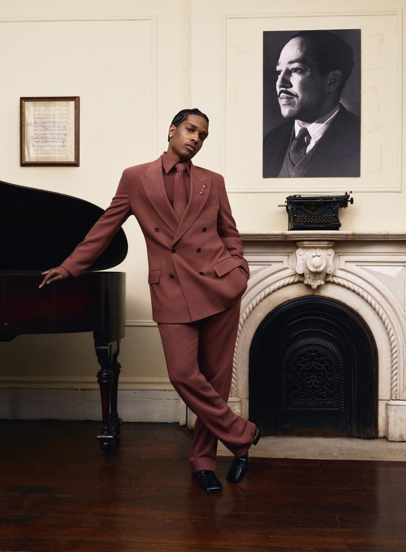 An adult male in a brown suit poses next to a grand piano in a room with an old fireplace and a framed portrait on the wall.