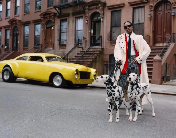 ASAP Rocky posing on a city street with two Dalmatians next to a yellow vintage car in front of brownstone buildings.
