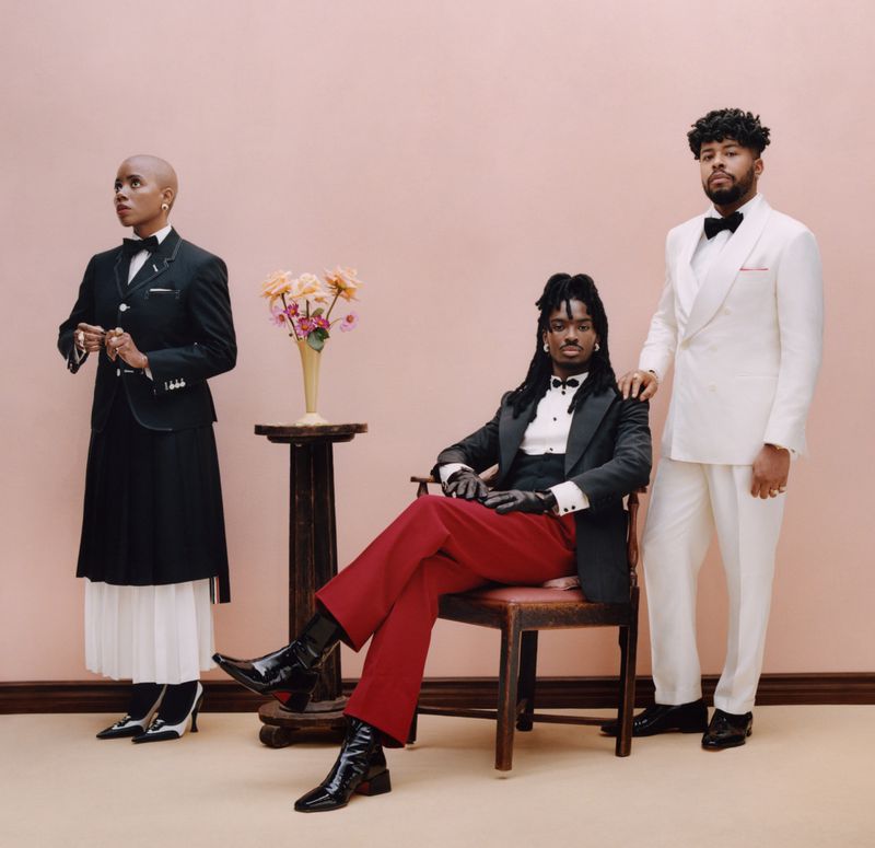 Three people in formalwear pose in a studio: a standing bald adult female, a seated adult with dreadlocks, and a standing adult male.