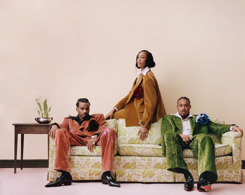 Three fashionable individuals in velvet suits pose indoors on a floral couch in front of a peach-colored wall with a minimalist side table.