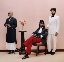 Three people in formal tailoring pose against a muted pink wall with a small vase of flowers on a pedestal table.