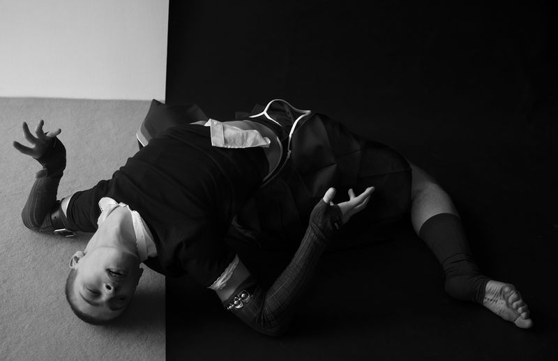Monochrome studio portrait of a person lying down in dark clothing, featuring striking contrast and dramatic lighting.