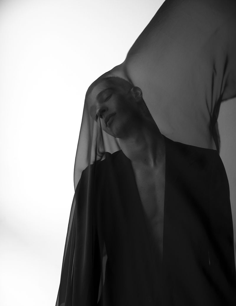 Monochrome portrait of a person with closed eyes, draped in sheer fabric, against a bright background, conveying an ethereal mood.