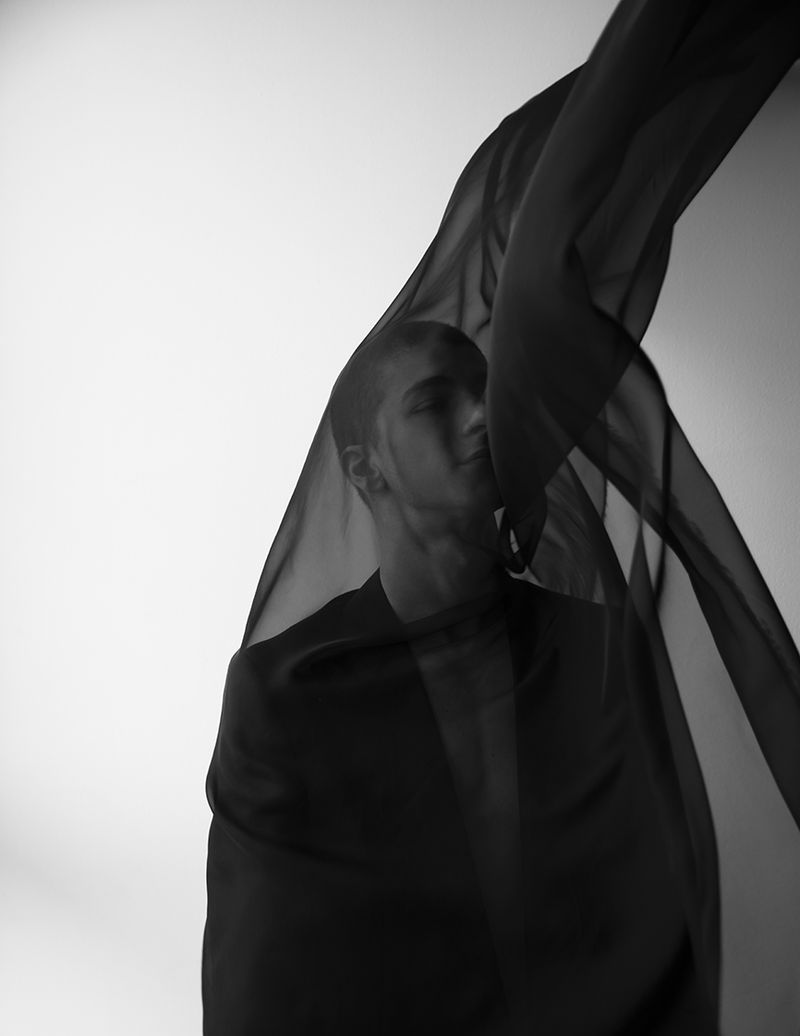 Black and white portrait of a young adult man partially obscured by a draped sheer black fabric against a plain light background.