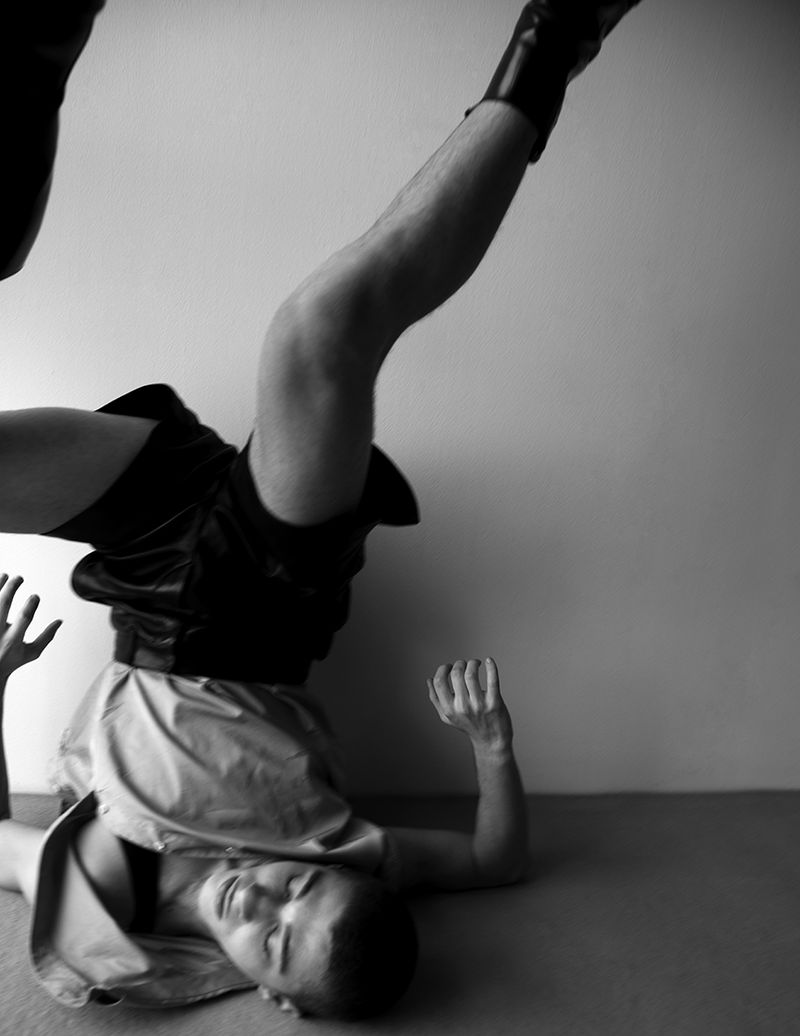 Black and white portrait of an adult male doing a headstand against a light wall wearing shorts and boots in an indoor setting.