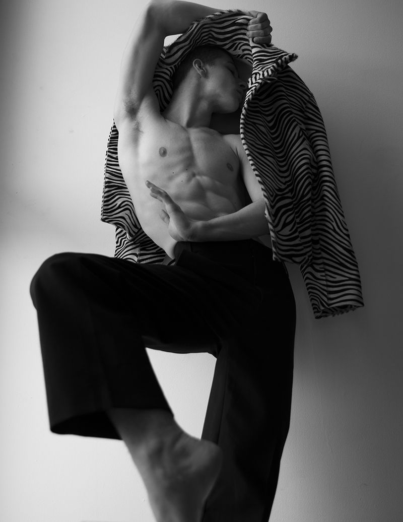 Striking black-and-white portrait of a muscular man in black pants, partially draped with a zebra-striped jacket, posing indoors.
