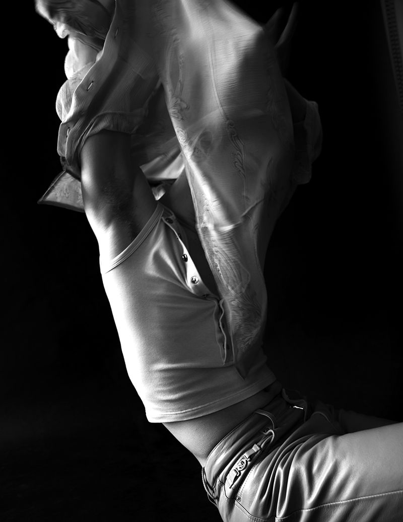 Black and white photo of an adult putting on a blouse with buttoned tank top and pants against dark background.